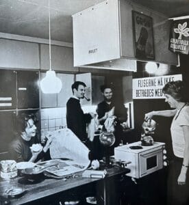 socialtmøde i køkkent hvor en kollegianer læser avis, en anden laver og the og de øvrige vasker op